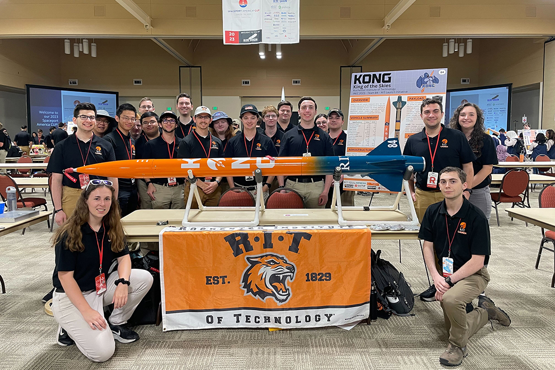 19 team members stand with orange rocket and RIT flag. 
