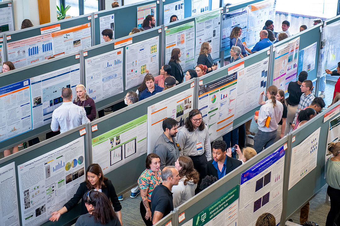 Students and faculty gather between four rows of research posters. People are walking through each row, asking researchers questions, and getting a close look at each poster.