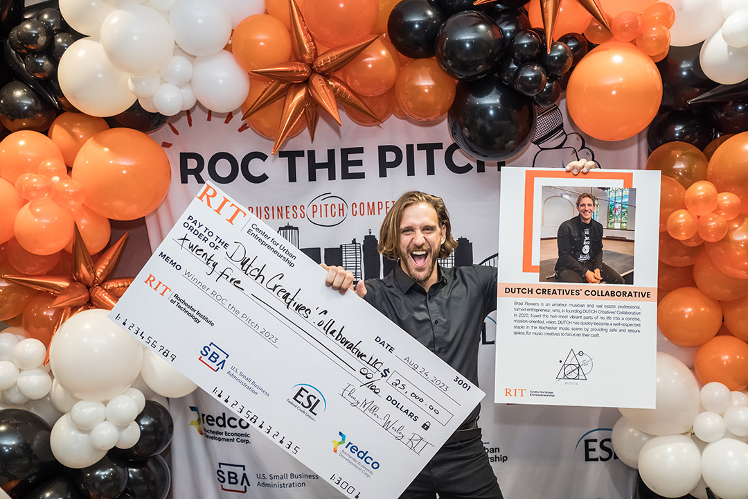 person surrounded by black, orange, and white balloons holding a giant novelty check for 25 thousand dollars and a poster with his photo and business name, Dutch Creatives Collaboration.
