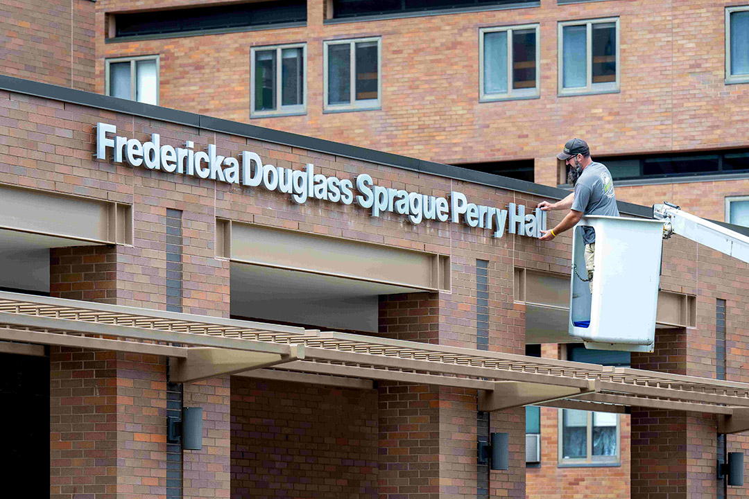 person in a cherry picker installing the last letter of Fredericka Douglass Sprague Perry Hall on the outside of a brick building.