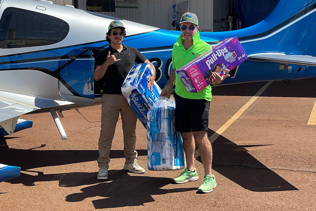 college student and father stand in front of a small airplane holding bags and boxes of bath tissue and diapers.