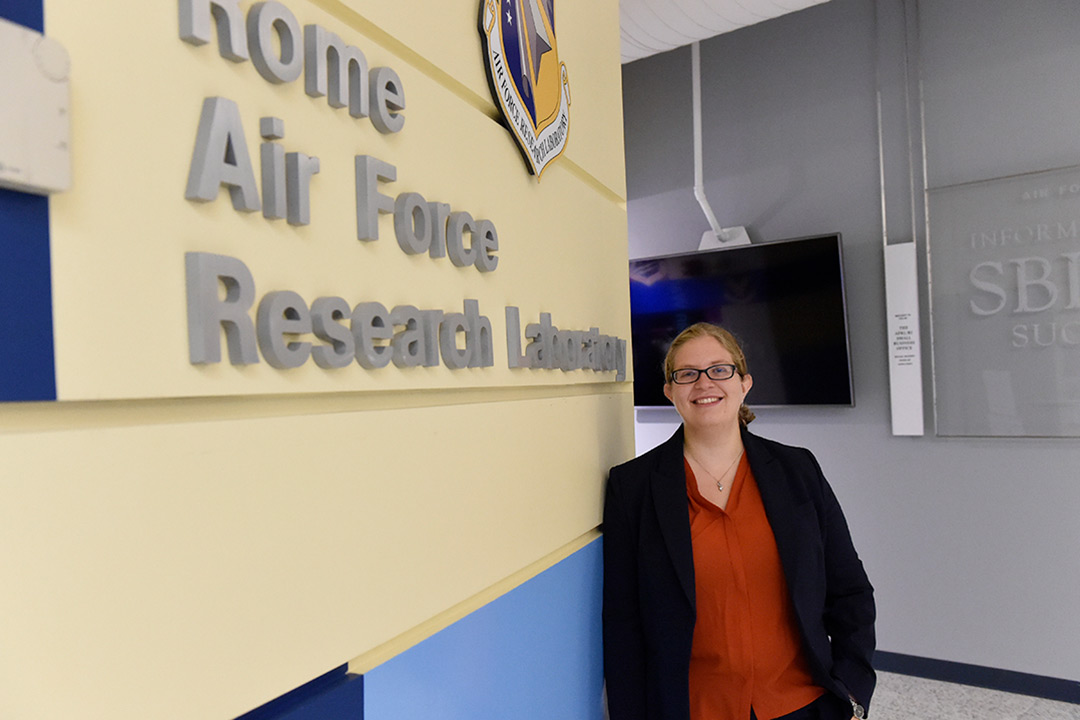 Karen Roth standing next to a sign that reads Rome Air Force Research Laboratory. She is smiling and wearing a red shirt and black blazer.