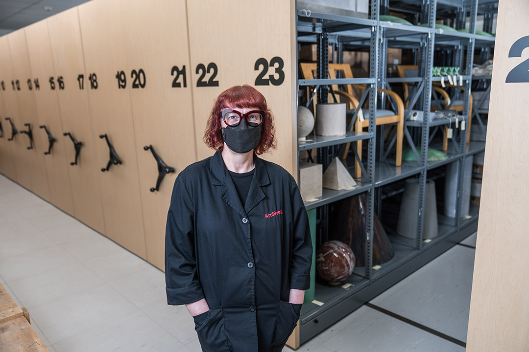 woman standing in front of a series of shelves wearing a black mask and black lab coat.