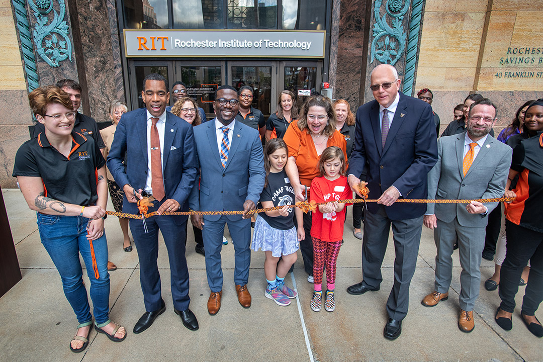 a group of people, including R I T and city officials as well as children, cut a ceremonial ribbon in front of a historic building.