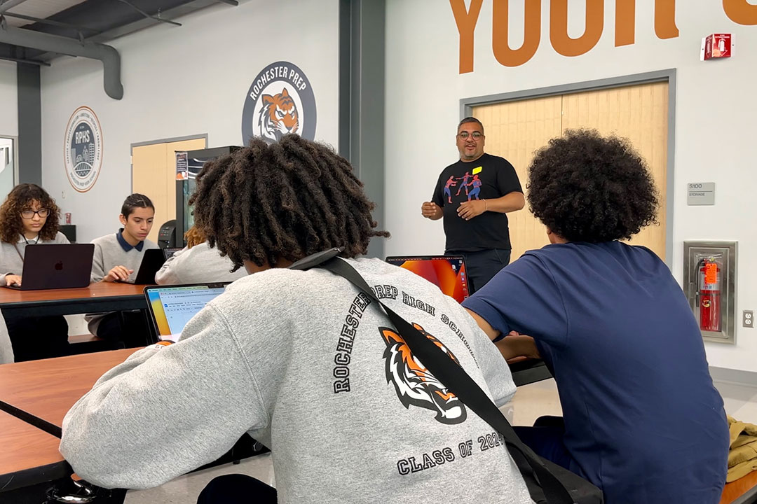 Man wearing spider-man shirt talks with high school students who are taking notes on a computer