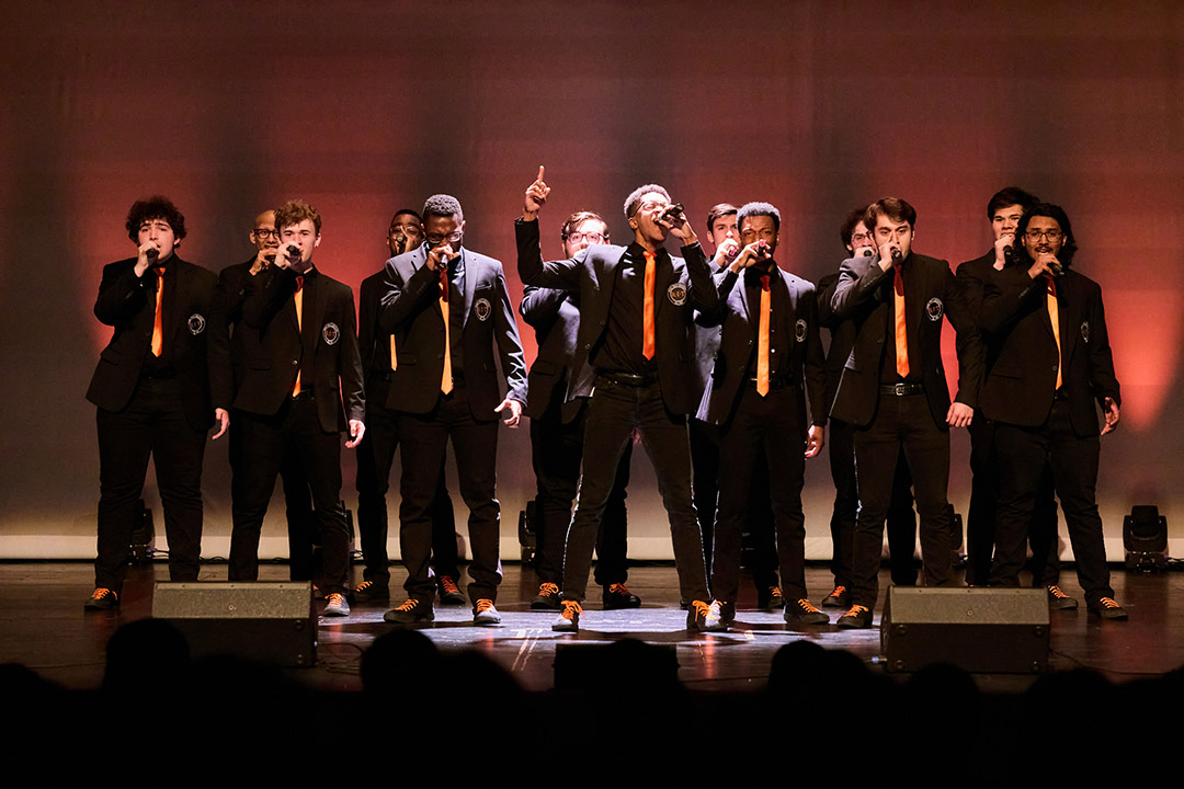 13 male college students wearing black suits lined up on a stage, singing.