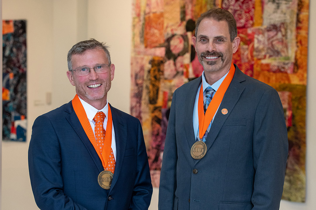 two college professors standing next to each other, both wearing R I T medallions around their necks.