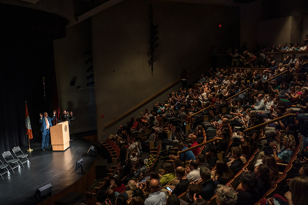 a full auditorium with the president of R I T on stage shaking his hands in the air for the American sign language sign for applause.