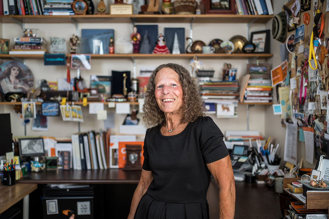 woman standing in her office, full of shelved of photos and knickknacks. 