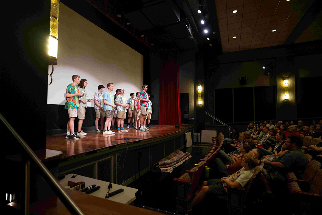 college students wearing Hawaiian shirts standing on a stage in a theater in front of a full crowd.