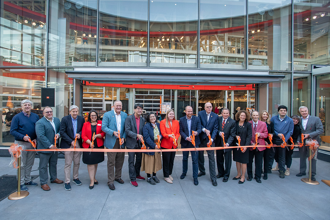 17 people lined up holding giant pairs of scissors in front of an orange ribbon.