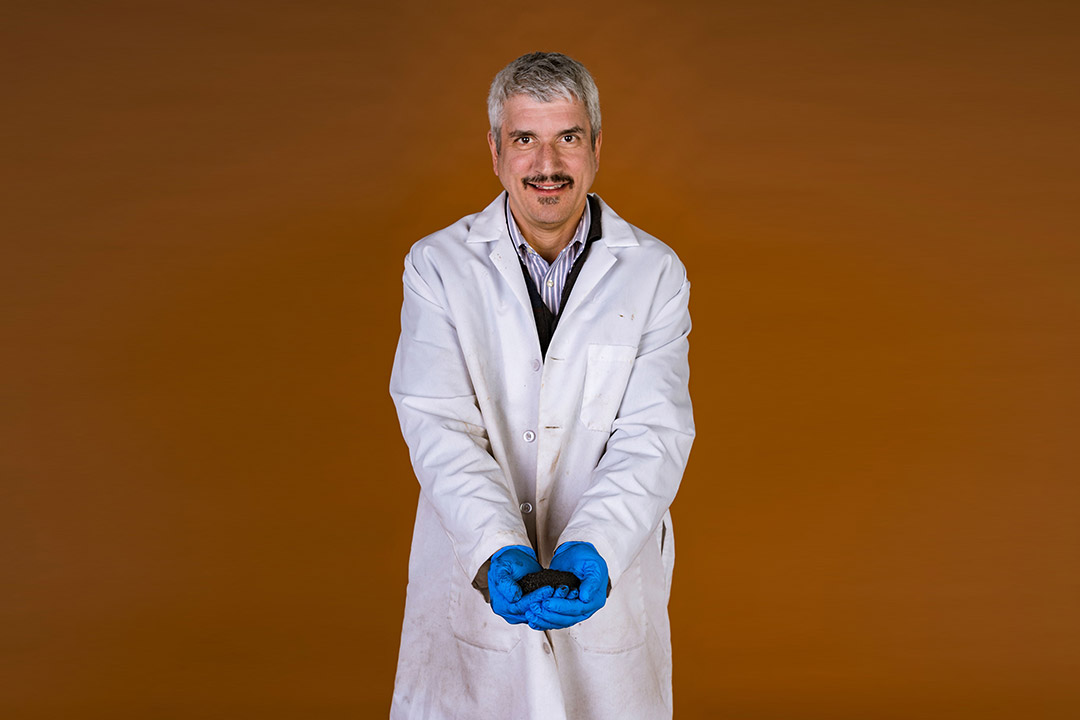 researcher wearing a white lab coat and blue rubber gloves holding what looks like black dirt.