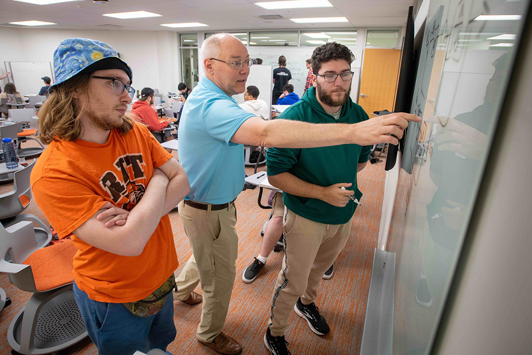 two college students standing next to a professor as he points to a dry erase board.