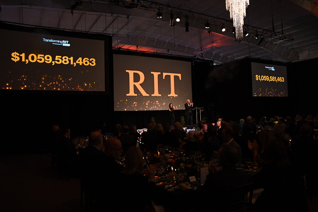 two people standing on a stage clapping with large projector screens behind them show the dollar amount of 1 billion, 59 million, 5 hundred 81 thousand, 4 hundred 63.