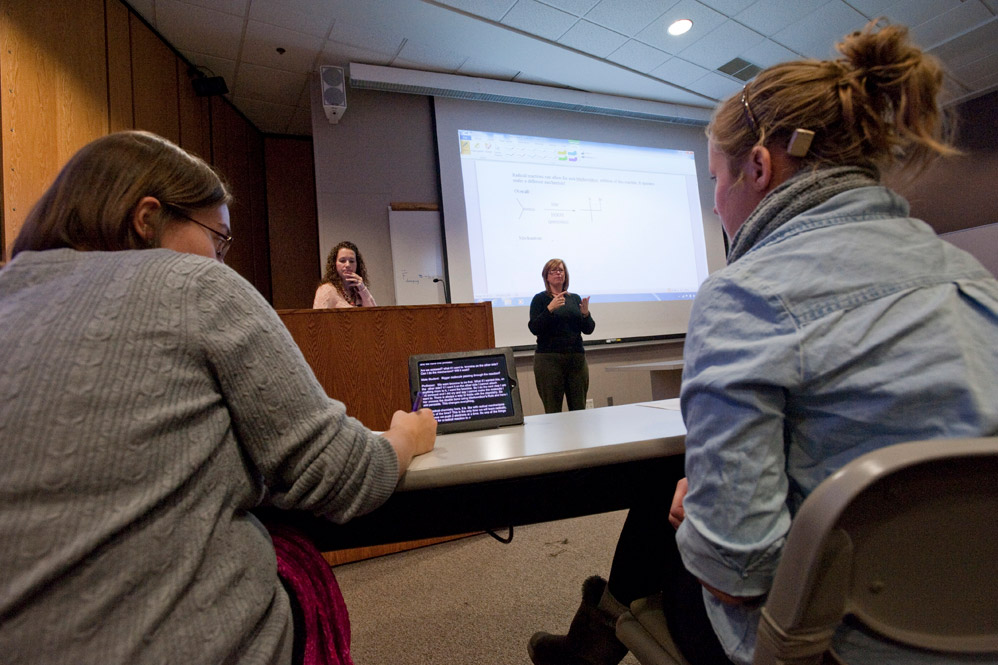 two college students in a classroom reading text on a tablet as a professor and A S L interpreter stand at the front of the room.