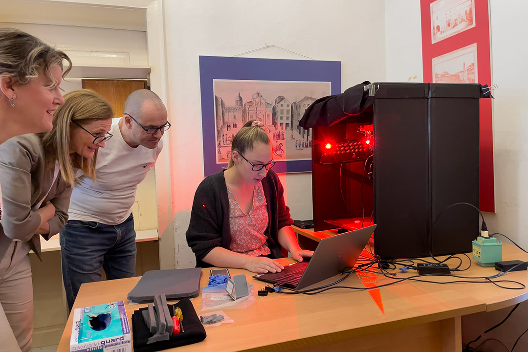 college student seated next to a covered box with red lights as three faculty watch her work on a laptop.