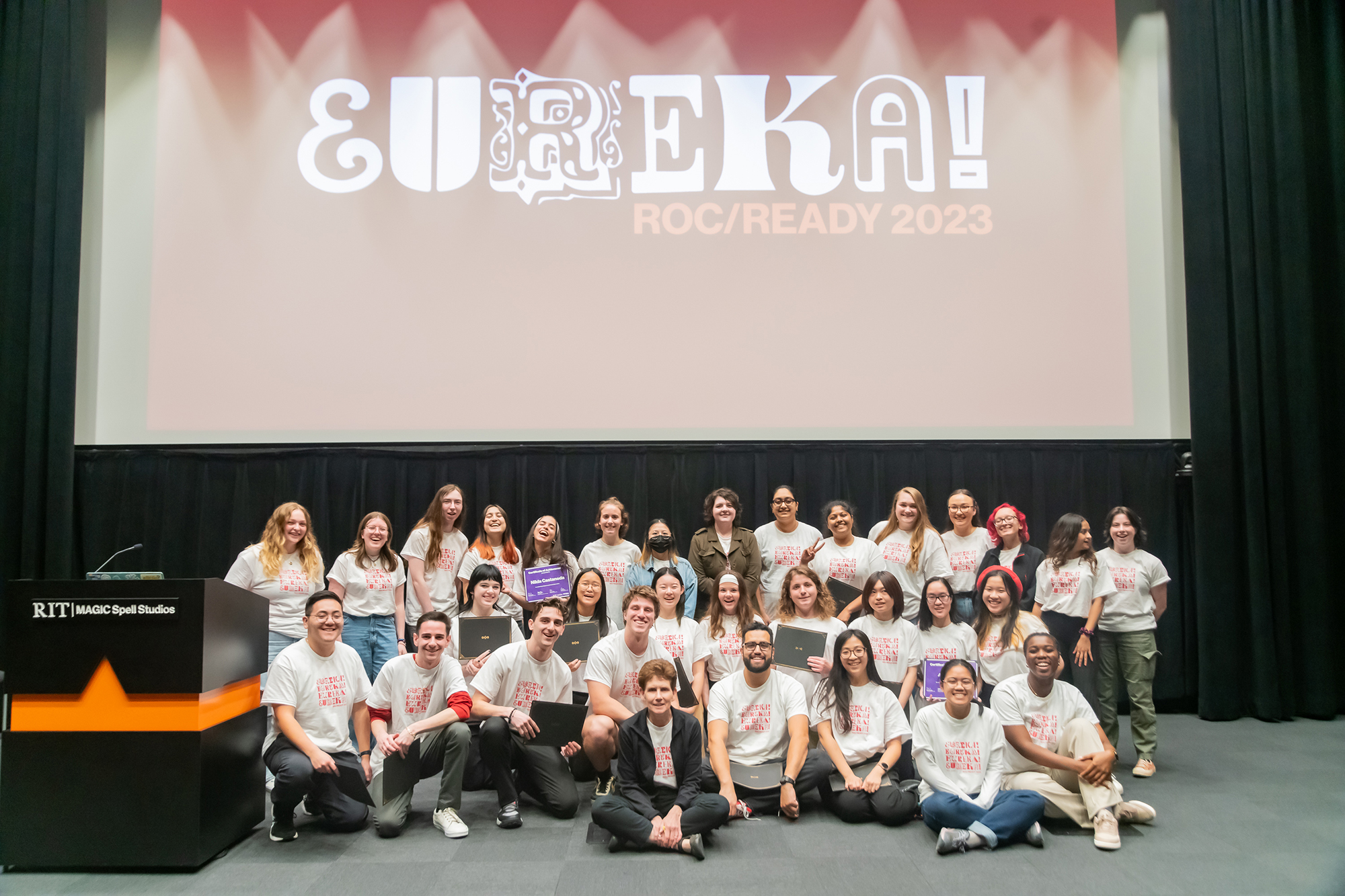 A group of EUREKA! 2023 participants with a projected program logo behind them.,