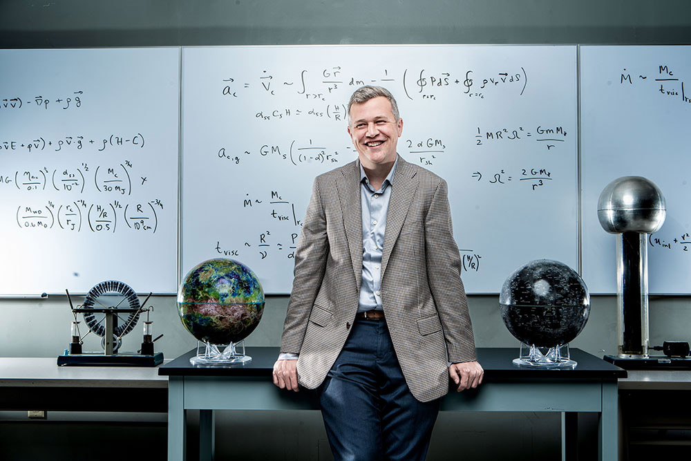 professor sitting on desk with globes on either side of him and whiteboards with math equations behind him.