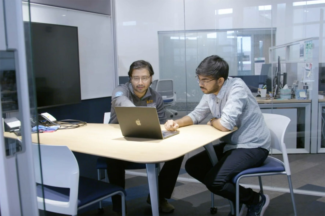 Ashique and Sujan sit at table in research lab pointing at laptop.