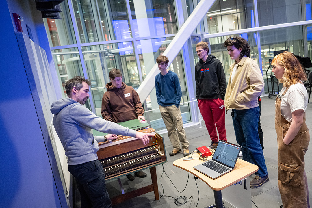 five college students standing around their professor, who is pointing to a laptop and adjusting a harpsichord.