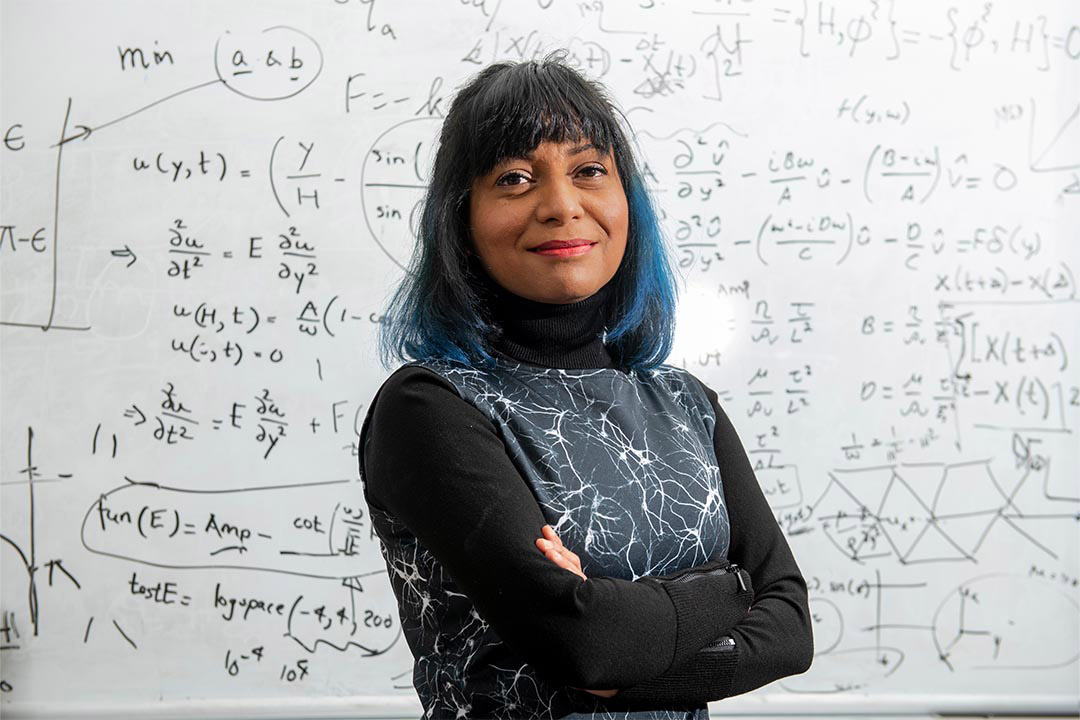 college professor posing with her arms crossed in front of a whiteboard covered in math equations.