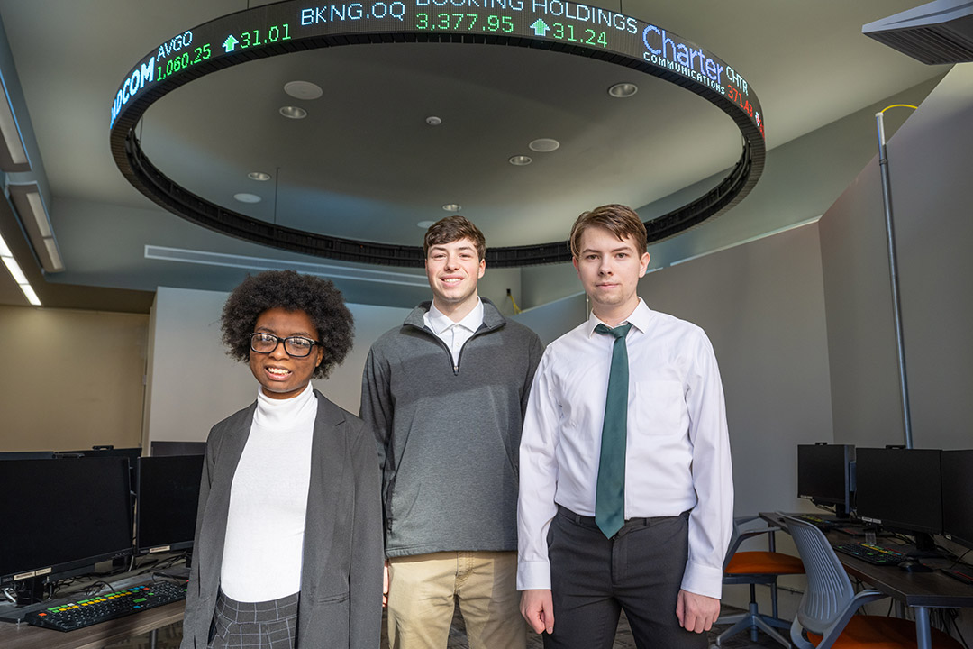 three college students standing in a lab with a lighted ribbon marquee sign above them showing real time stock points.
