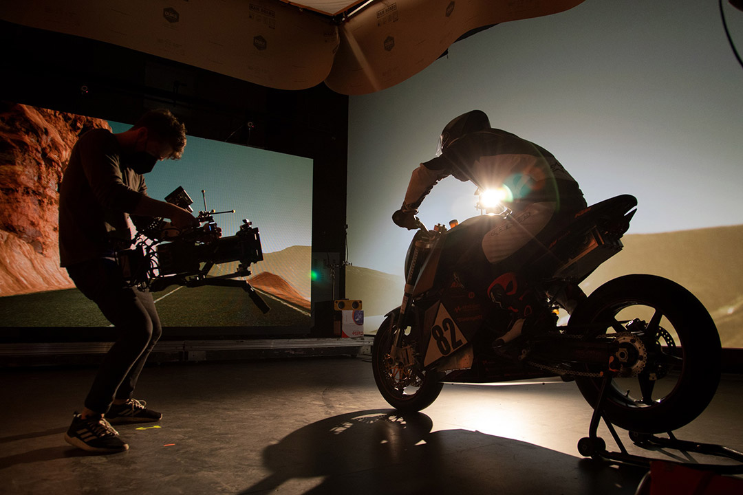 college student holding a steady cam pointed at a person on a motorcycle inside a film studio.