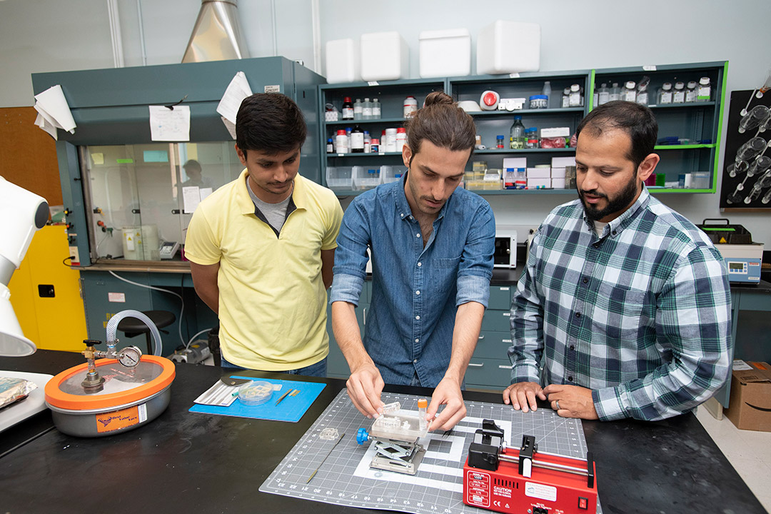 two college students and a professor test cell movement on a tiny scissor lift in a lab.