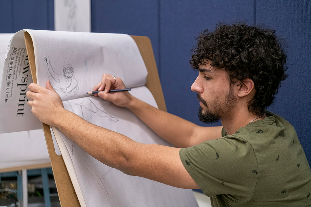 a student in a green shirt sketching on a large pad of paper that is sitting on an easel.