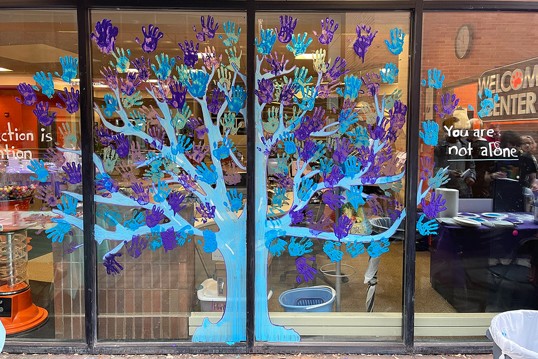 glass windows with a blue tree painted on it. There are blue and purple handprints to make up the leaves. Text on the window says you are not alone.