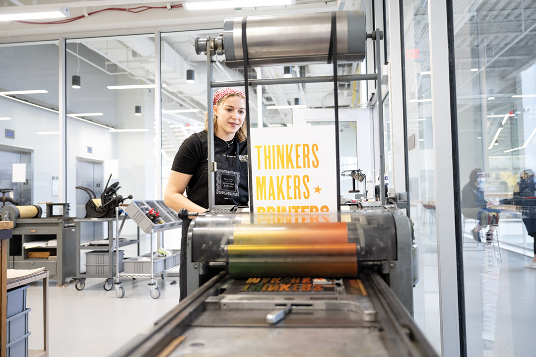 college student runs a vintage printing press, making a poster that says thinkers, makers, printers in gold ink.