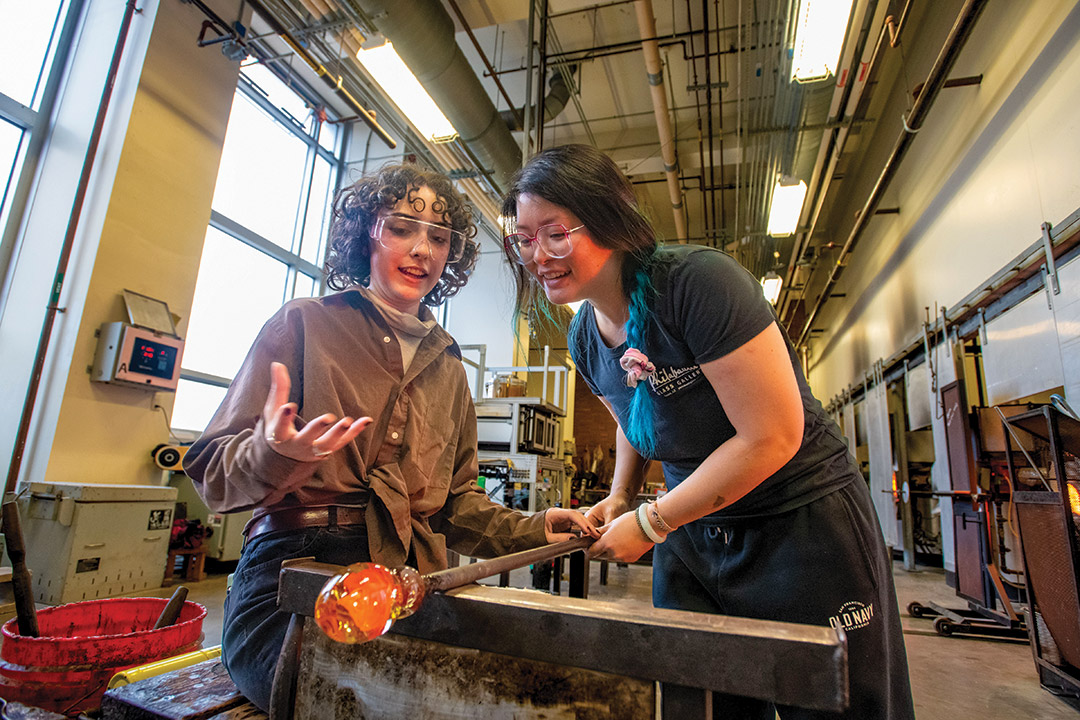 artist guiding a college student using a pole to rotate molten glass in a hot shop.