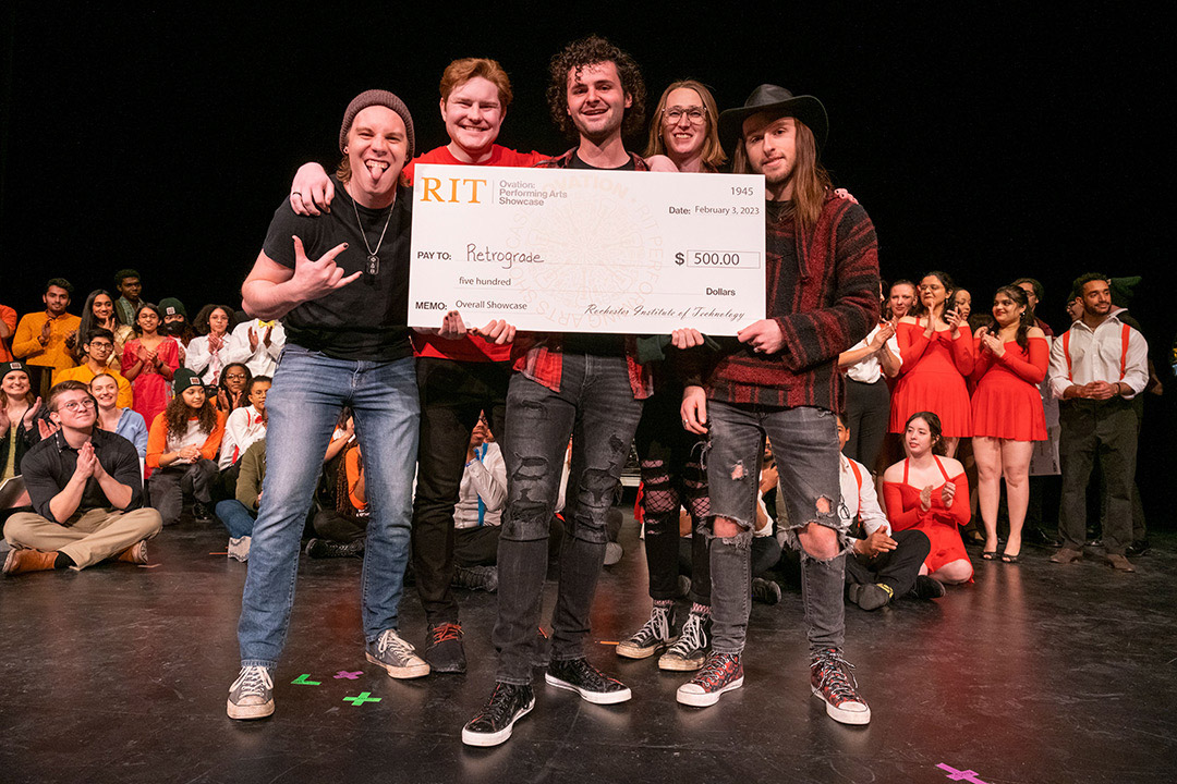 Five members of the winning band stand on stage holding a giant check from RIT that notes their 500-dollar prize. Behind them is a small crowd of other performers.