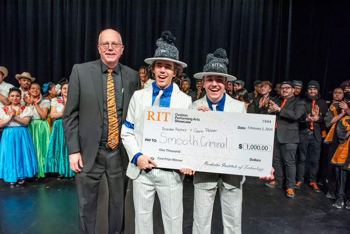David Munson, Jr., RIT President, stands on stage with 2 students that won the $1,000 prize. Other performers stand behind them