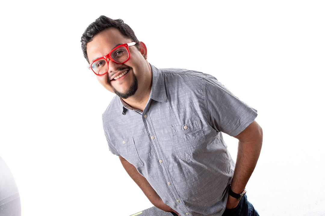 Juan Noguera is shown standing with a short sleeve chambray shirt and red framed glasses