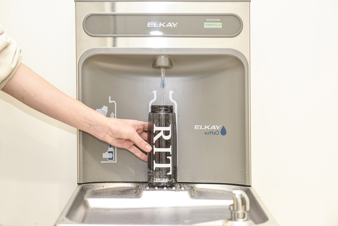 a water fountain is shown with a retrofitted nozzle to fill water bottles and a hand is holding a water bottle with the letters RIT on it under the stream of water