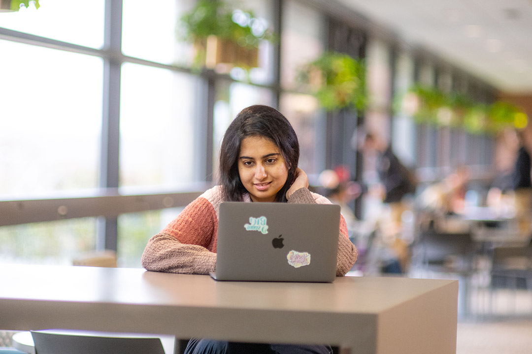 a student is shown sitting at a table in the atrium of a building working on a laptop.