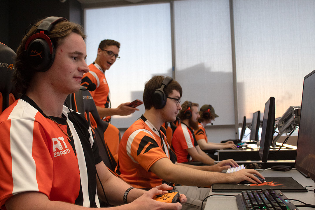 The RIT Esports Halo team practices for competition sitting at a desk with computers wearing orange and white team shirts.