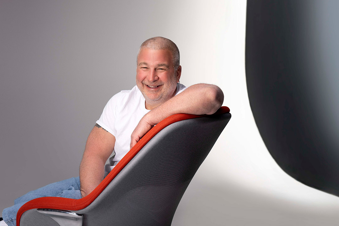 Carl Salvaggio is shown sitting on a gray and orange chair with his arm resting casually on the top of the chair.