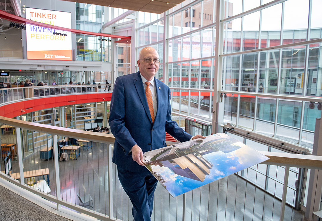 David Munson, Jr., RIT President, stands on the second floor balcony of the SHED holding a portrait that shows the renderings before it was built. The.