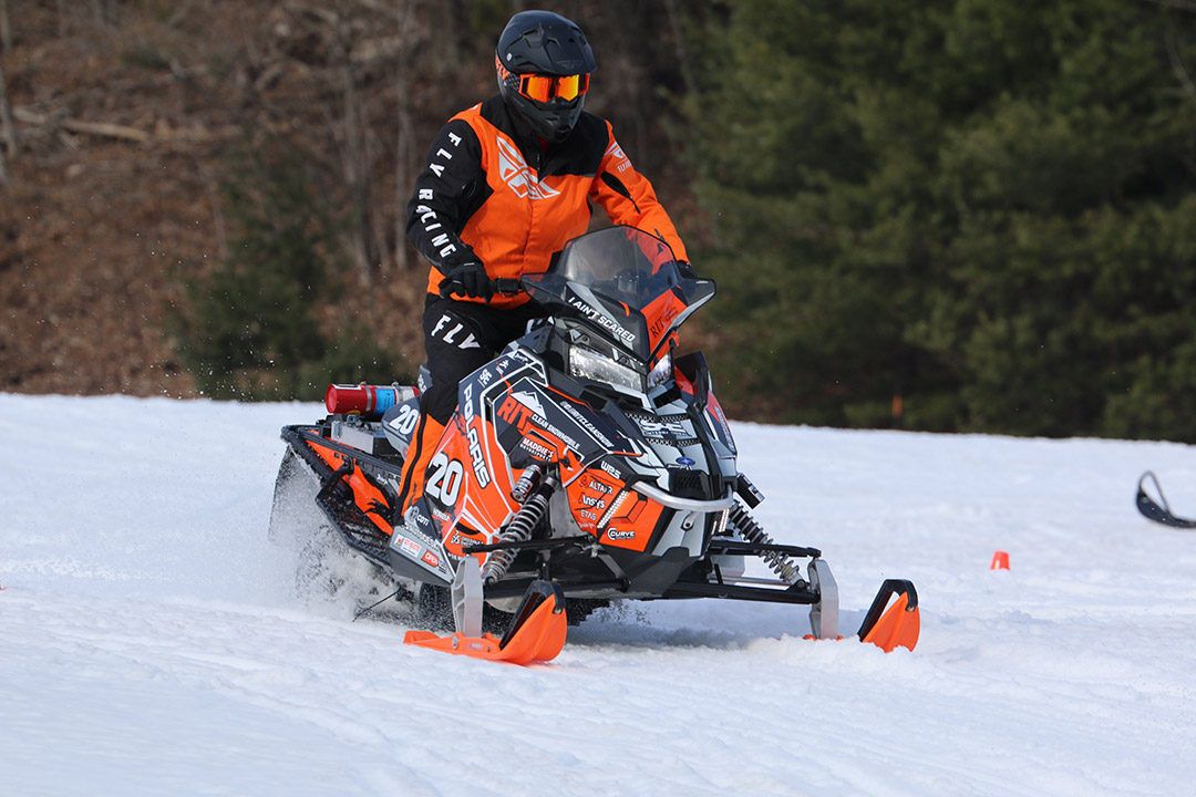 a student is shown in an orange snowsuit riding a snowmobile through snow.