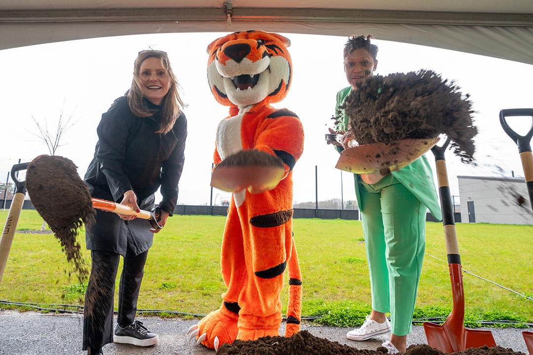 Sandra Johnson, Ritchie the Tiger, and Jackie Nicholson stand under a tent with shovels that are covered in dirt signifying a groundbreaking.