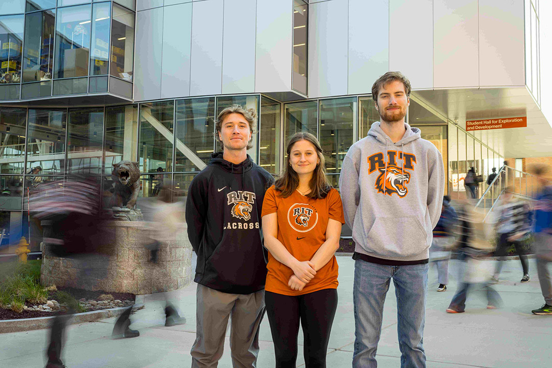 Three students stand in front of the SHED on campus, all in R I T shirts.