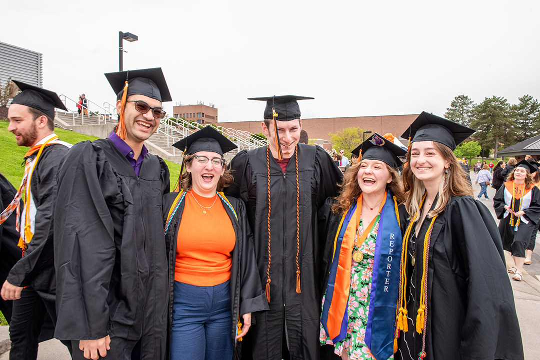 'Five graduates stand next to each other in regalia in front of the Gordon Field House.'