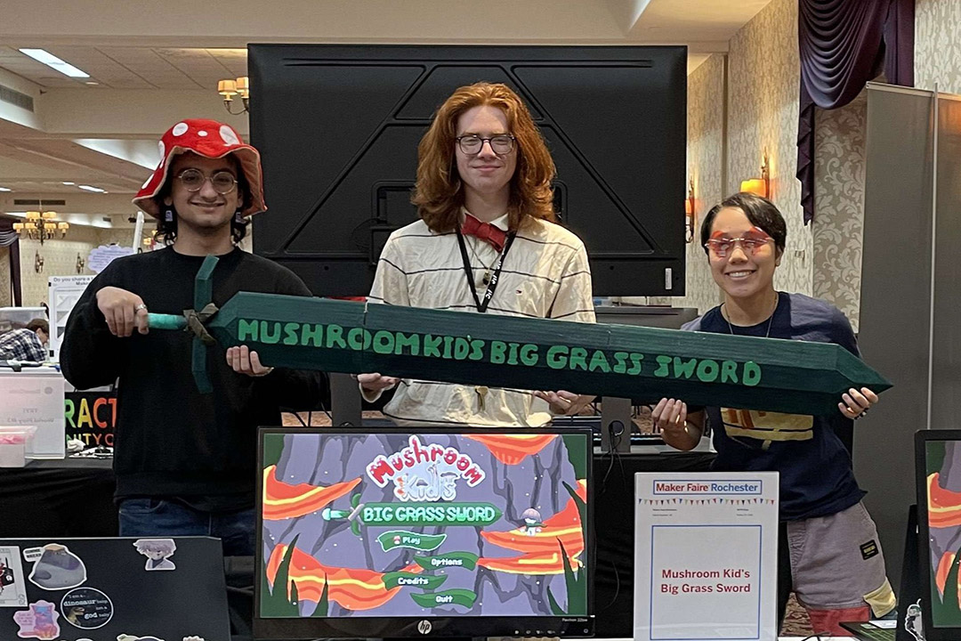 Three students stand in front of a monitor with their game displayed holding a sign with the game name.