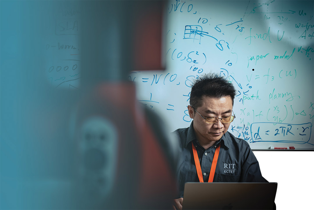 a man looks at a computer laptop screen while sitting in front of a whiteboard with formulas scribbled across it.