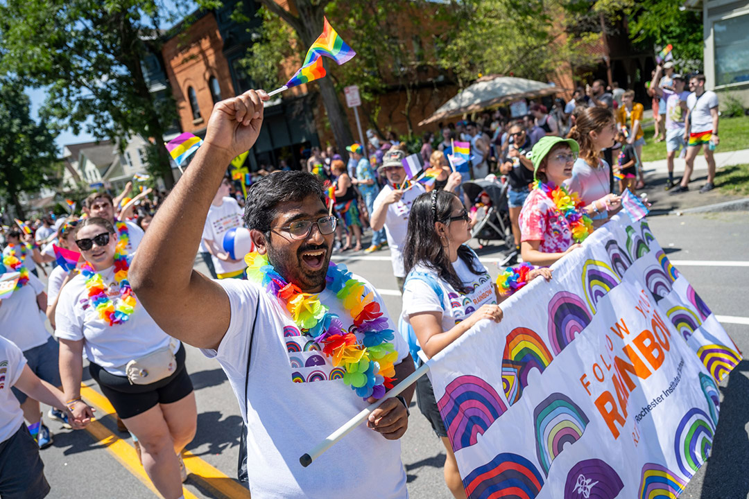 Many people walk on the street at a parade wearing rainbow items and waving rainbow flags.