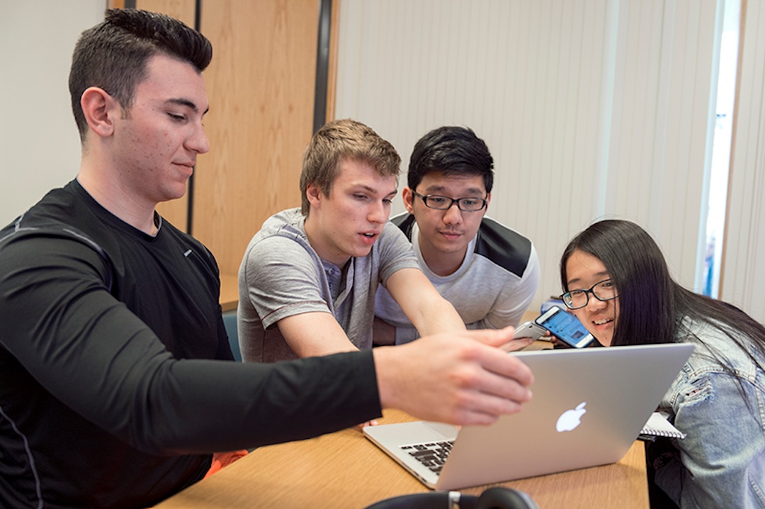 4 students sit around a table looking together at the screen of a single MacBook