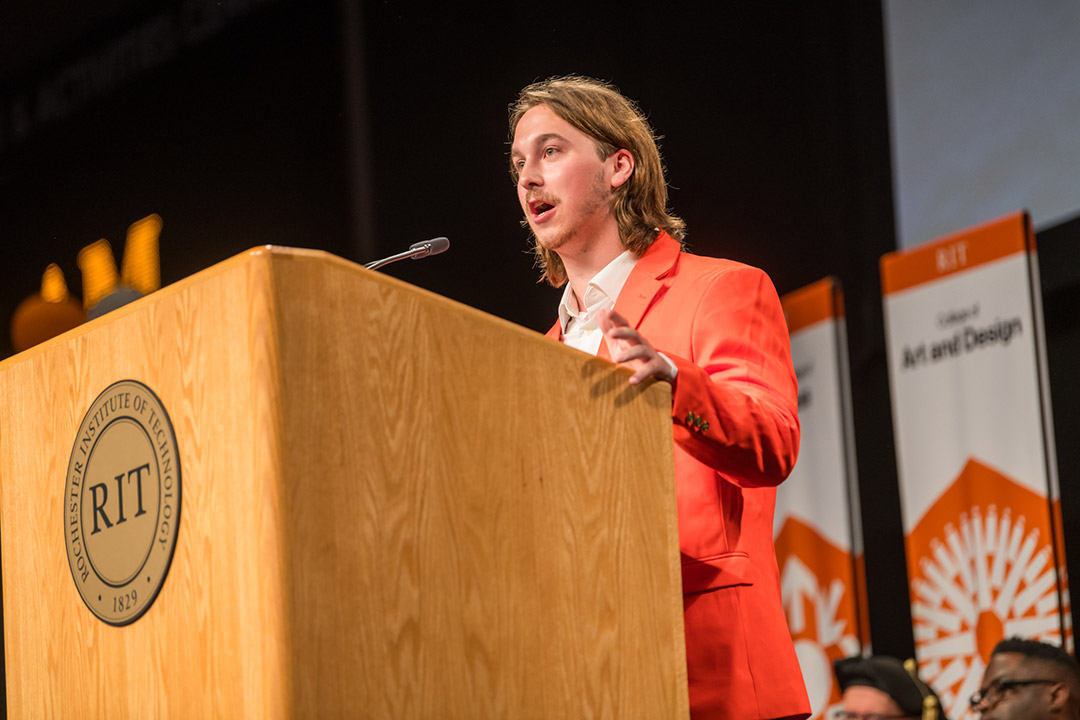 Alex Shuron stands at a podium addressing students at convocation.