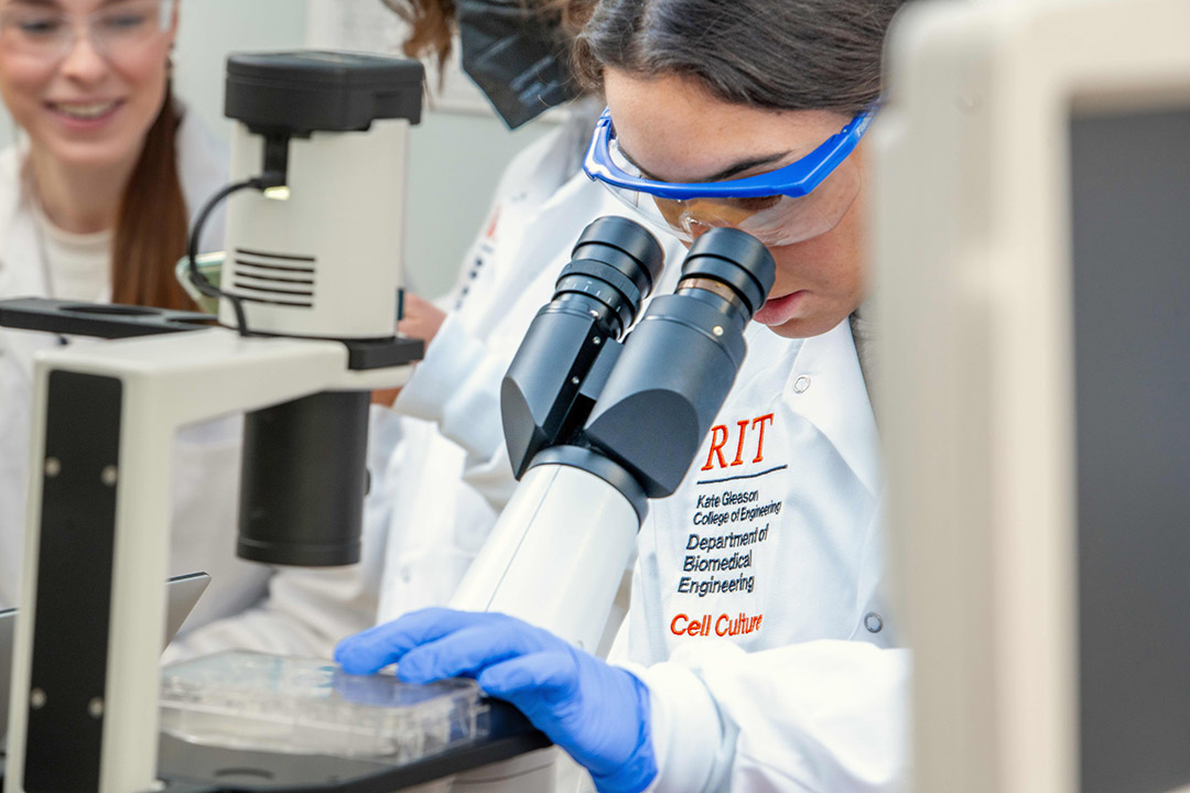 a student in a white lab coat uses a microscope to look at cells in a lab.